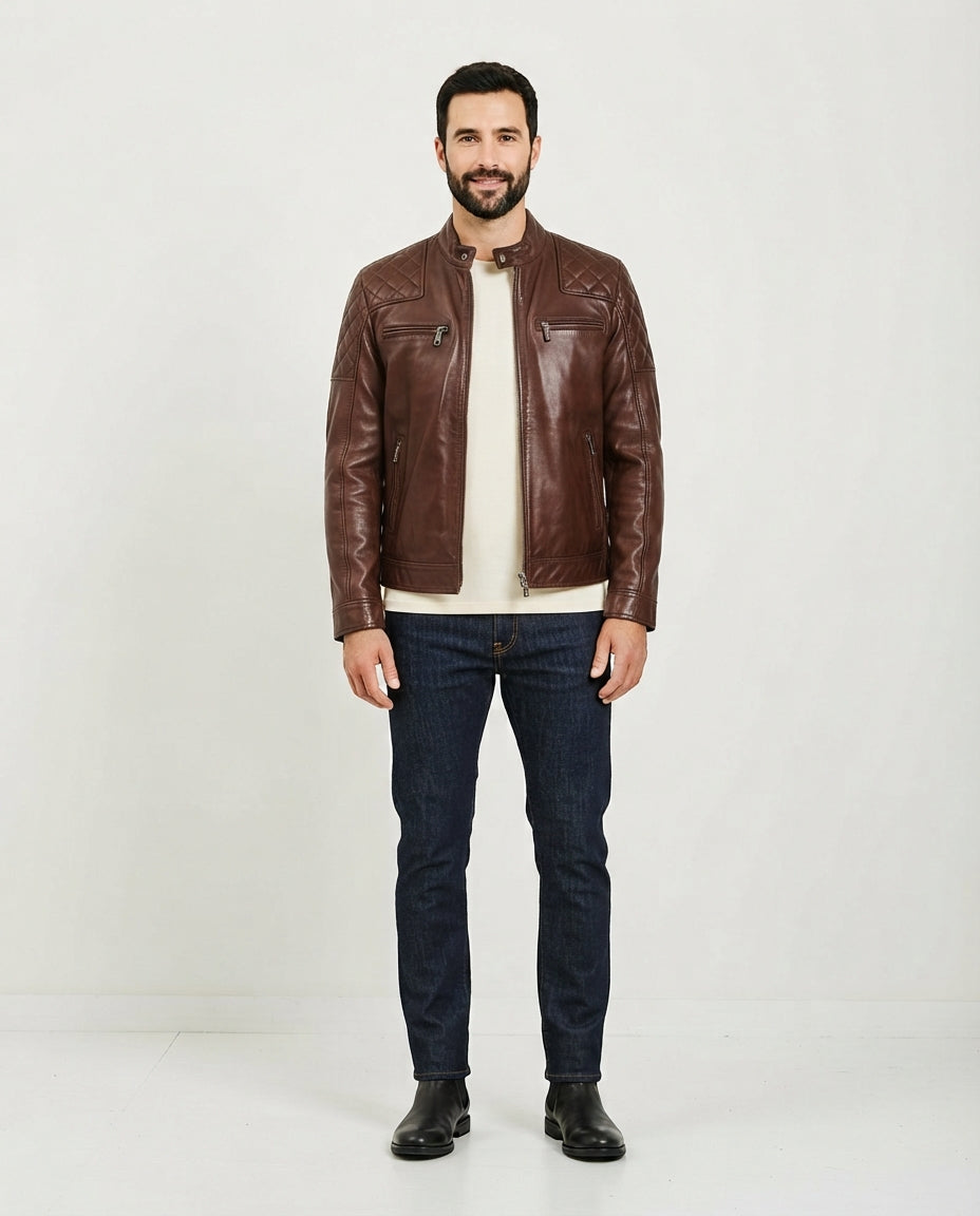 A man wearing a brown leather jacket over a cream t-shirt, dark blue jeans, and black boots, standing against a plain white studio background.
