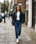 A woman walking on a city sidewalk wearing a black leather moto jacket, white t-shirt, blue jeans, and white sneakers.