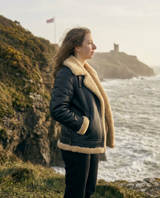 A woman viewed from behind wearing a Black shearling-lined jacket, standing on a rocky cliffside overlooking the ocean with a castle in the distance.
