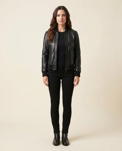 A woman stands against a plain beige studio background wearing a black leather bomber jacket over a black top and black skinny pants with black ankle boots.