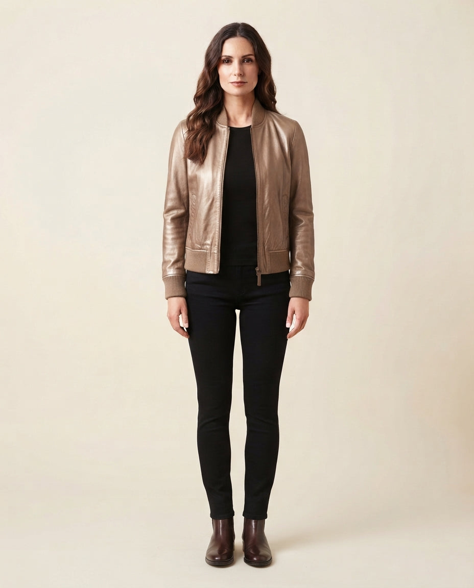 A woman stands against a plain beige studio background wearing a metallic taupe leather bomber jacket over a black top and black skinny pants, with dark brown ankle boots.