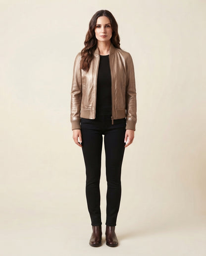 A woman stands against a plain beige studio background wearing a metallic taupe leather bomber jacket over a black top and black skinny pants, with dark brown ankle boots.