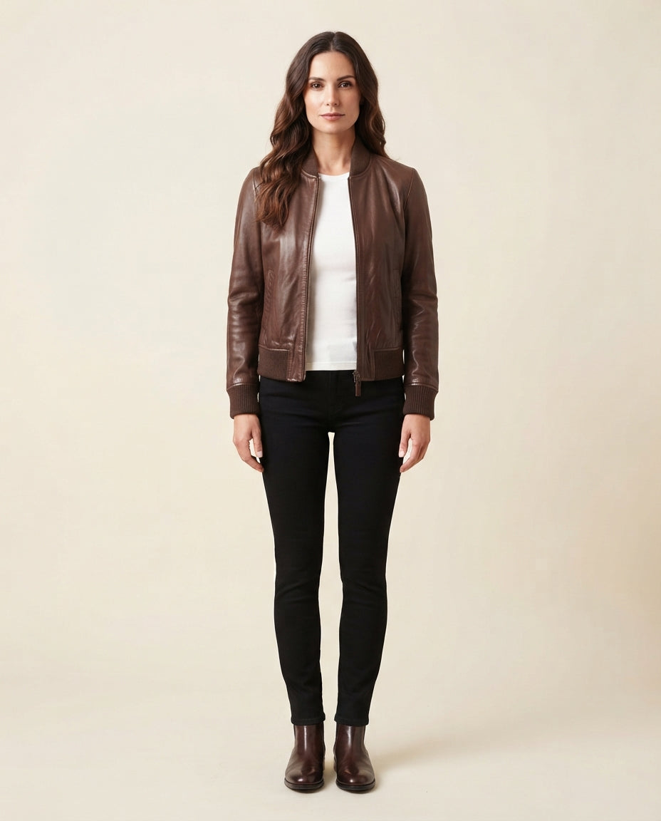 A woman stands against a plain beige studio background wearing a brown leather bomber jacket over a white top, black skinny pants, and dark brown ankle boots.