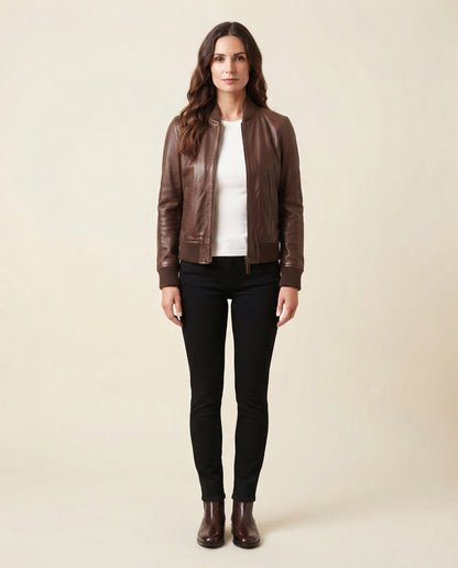 A woman stands against a plain beige studio background wearing a brown leather bomber jacket over a white top, black skinny pants, and dark brown ankle boots.