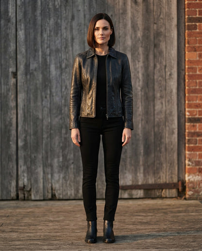 A woman stands outdoors in front of a weathered wooden door and brick wall, wearing a black leather moto jacket over a black top and black skinny pants.