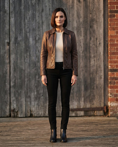 A woman stands outdoors in front of a weathered wooden door and brick wall, wearing a brown leather moto jacket over a cream top with black pants and black ankle boots.