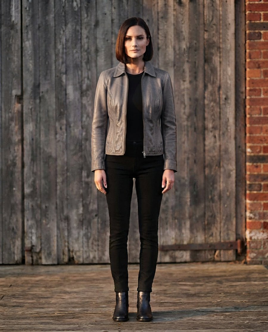 A woman stands outdoors in front of a weathered wooden wall and brick column, wearing a fitted light gray leather jacket over a black top and black skinny pants with black ankle boots.
