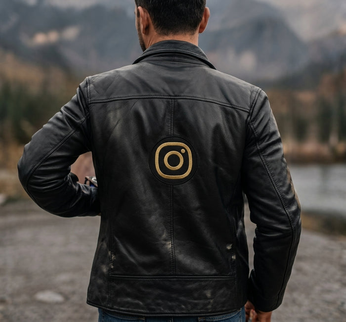 A man viewed from behind wearing a black leather jacket with a gold Instagram logo printed on the center back, set against a blurred outdoor mountain and lake background.
