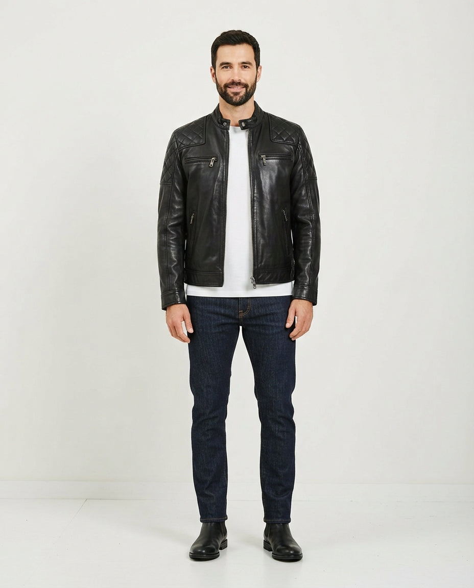 A man wearing a black leather biker jacket over a white t-shirt, dark blue jeans, and black boots, standing against a plain white studio background.