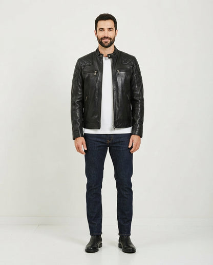 A man wearing a black leather biker jacket over a white t-shirt, dark blue jeans, and black boots, standing against a plain white studio background.