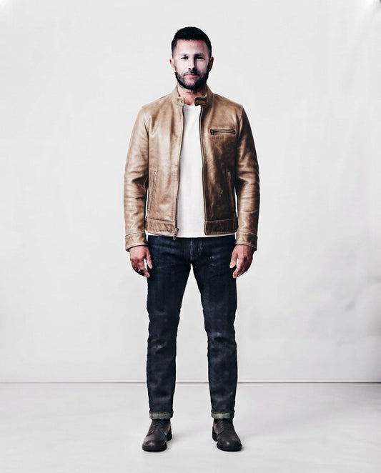A man wearing a light brown leather jacket over a cream t-shirt, dark blue jeans, and brown boots, standing against a plain white studio background.