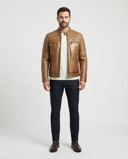 A man modeling a tan quilted leather jacket over a cream t-shirt, dark blue jeans, and brown leather boots, standing against a light gray studio background.