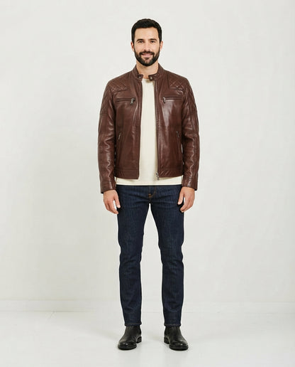 A man wearing a brown leather jacket over a cream t-shirt, dark blue jeans, and black boots, standing against a plain white studio background.