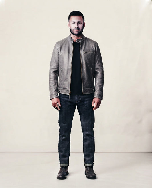 A man stands facing forward wearing a taupe-brown leather jacket over a black shirt, dark blue jeans, and dark boots, photographed against a plain light beige studio backdrop.