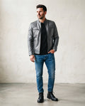 A man wearing a silver leather jacket with quilted shoulder details, a black t-shirt, blue jeans, and black boots, standing against a textured off-white wall.