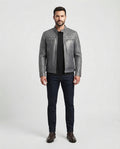A man modeling a gray quilted leather jacket over a black shirt and dark blue jeans, standing against a light gray studio background.