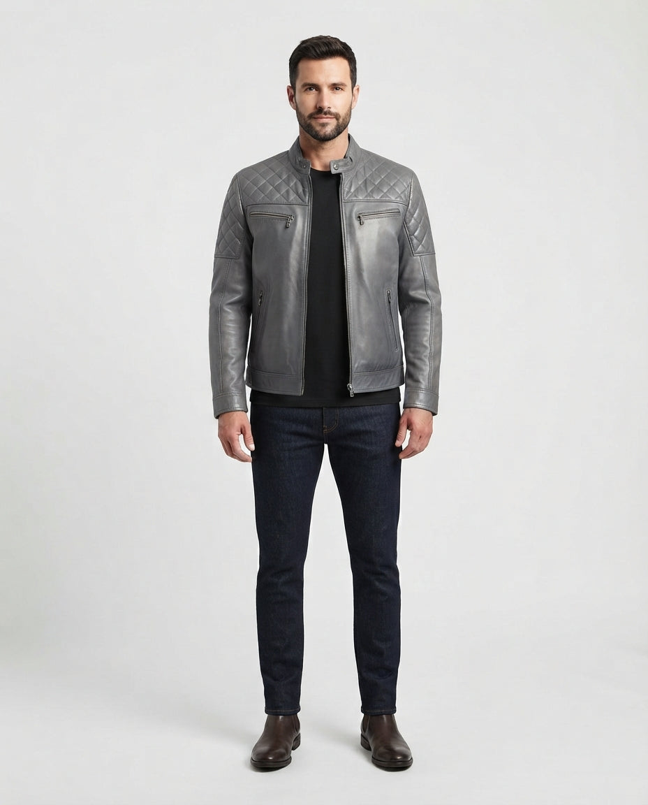 A man modeling a gray quilted leather jacket over a black shirt and dark blue jeans, standing against a light gray studio background.