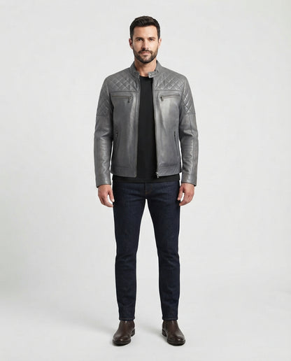 A man modeling a gray quilted leather jacket over a black shirt and dark blue jeans, standing against a light gray studio background.