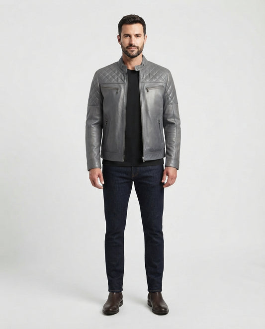 A man modeling a gray quilted leather jacket over a black shirt and dark blue jeans, standing against a light gray studio background.