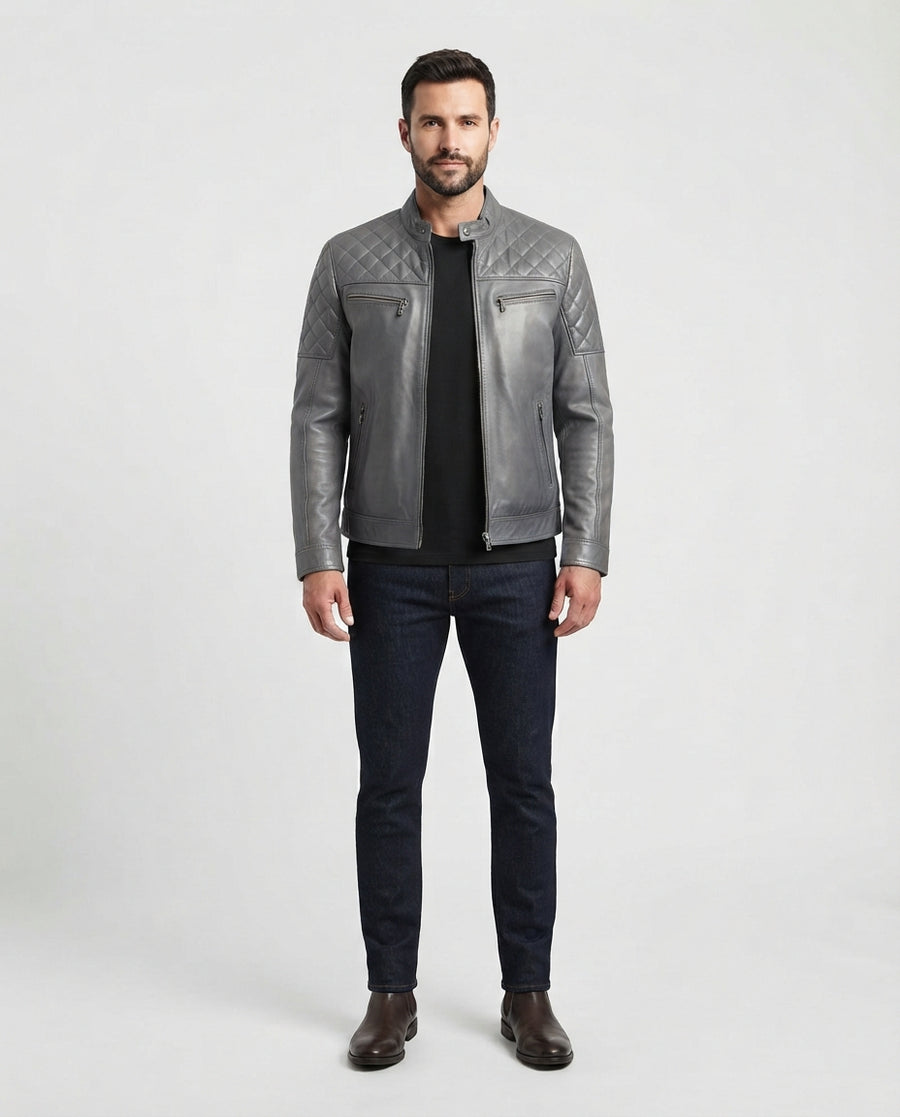 A man modeling a gray quilted leather jacket over a black shirt and dark blue jeans, standing against a light gray studio background.