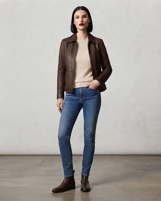 A woman stands in a studio wearing a dark brown leather jacket over a light beige top, blue skinny jeans, and dark brown ankle boots against a neutral light gray background.